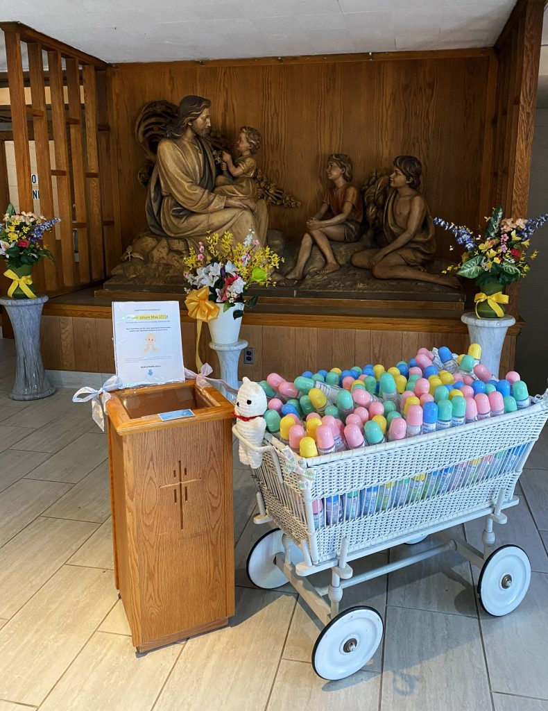 Church display featuring a religious statue of Jesus seated with children behind a donation stand and a white wagon filled with colorful baby bottles for a baby bottle drive.